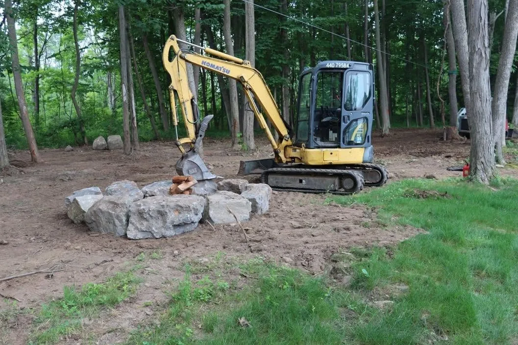A yellow Komatsu mini excavator in a wooded clearing, beside a ring of large boulders set around a fire pit