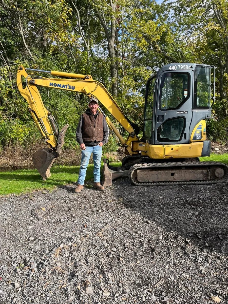 A man in a work vest and cap standing in front of a yellow Komatsu mini excavator on a gravel lot