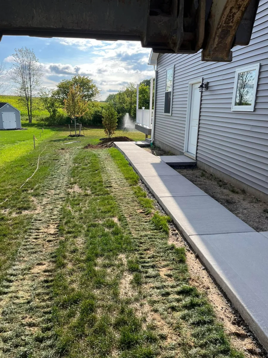 A new concrete walk along the side of a house, seen from under an excavator bucket, with the yard beside it seeded