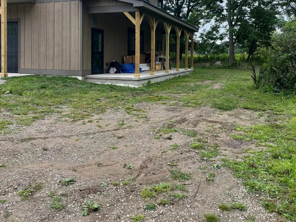 A rutted, weedy gravel yard beside a new barn-style house with a long covered porch